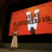 Žena v šatech stojí na podiu před plátnem promítajícím film