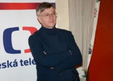 Muž v černém obleku stojí před plakátem s logem České televize