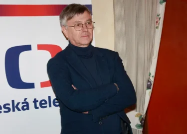 Muž v černém obleku stojí před plakátem s logem České televize