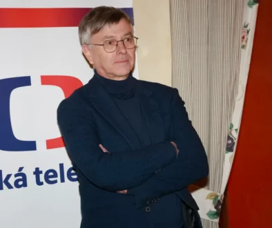 Muž v černém obleku stojí před plakátem s logem České televize