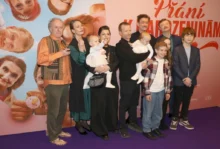 Skupina spokojených dospělých, menších i větších dětí před plakátem filmu Křtiny