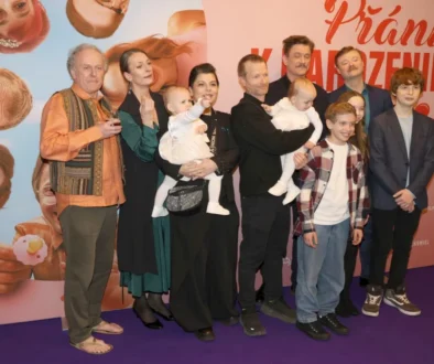Skupina spokojených dospělých, menších i větších dětí před plakátem filmu Křtiny