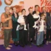 Skupina spokojených dospělých, menších i větších dětí před plakátem filmu Křtiny