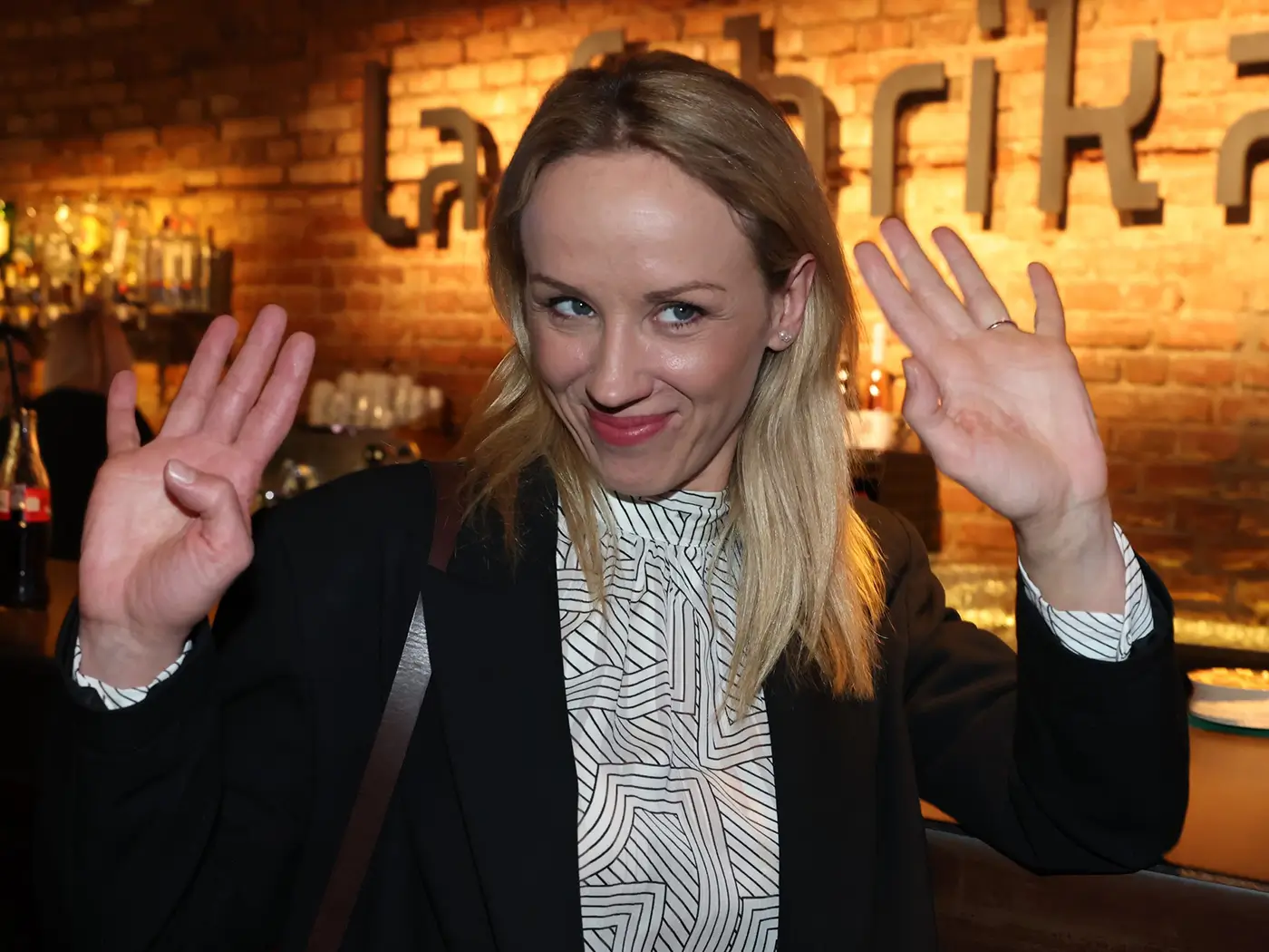 Žena v černém saku a se světlými vlasy zdvihá ruce nahoru
