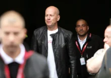 Muž v kožené bundě jde mezi bodyguardy. Má pleš a usmívá se