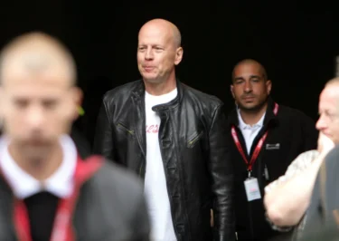 Muž v kožené bundě jde mezi bodyguardy. Má pleš a usmívá se