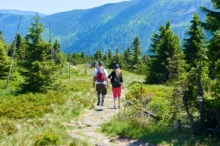 Dva turisté jsou pěšinou kolem se tyčí vysoké hory plné lesů