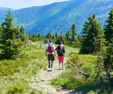 Dva turisté jsou pěšinou kolem se tyčí vysoké hory plné lesů