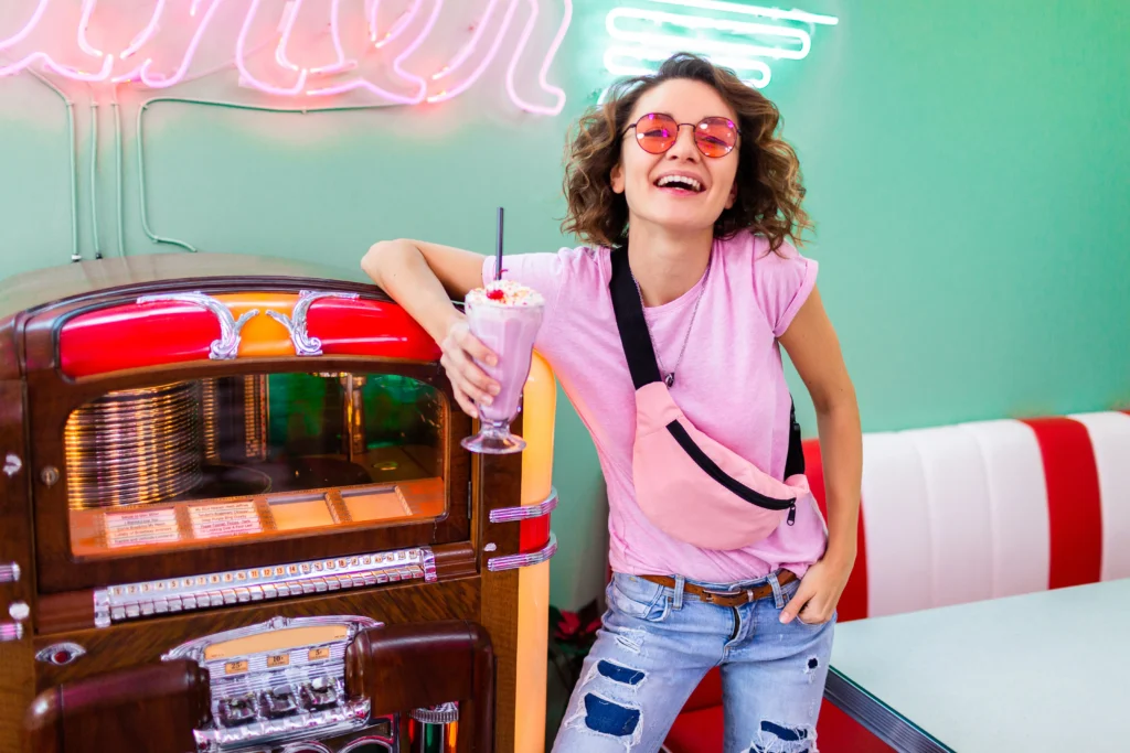 shutterstock_1354956533 - VIPshow Žena v růžovém tričku, džínách a s ledvinkou se opírá o klasický jukebox ve stylizaci 50. let minulého století