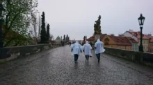 tři lidé v modrých pláštěnkách běží po kamenném mostě