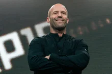 Smiling bald man on stage with folded arms, wearing a black button-up shirt, in front of a large LED screen.
