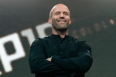 Smiling bald man on stage with folded arms, wearing a black button-up shirt, in front of a large LED screen.