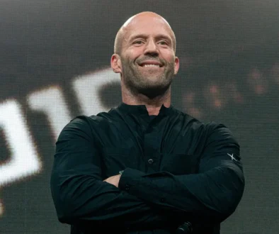 Smiling bald man on stage with folded arms, wearing a black button-up shirt, in front of a large LED screen.
