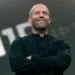 Smiling bald man on stage with folded arms, wearing a black button-up shirt, in front of a large LED screen.