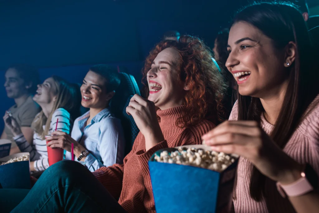 Group of friends in a dark cinema laughing together, with popcorn and drinks nearby.