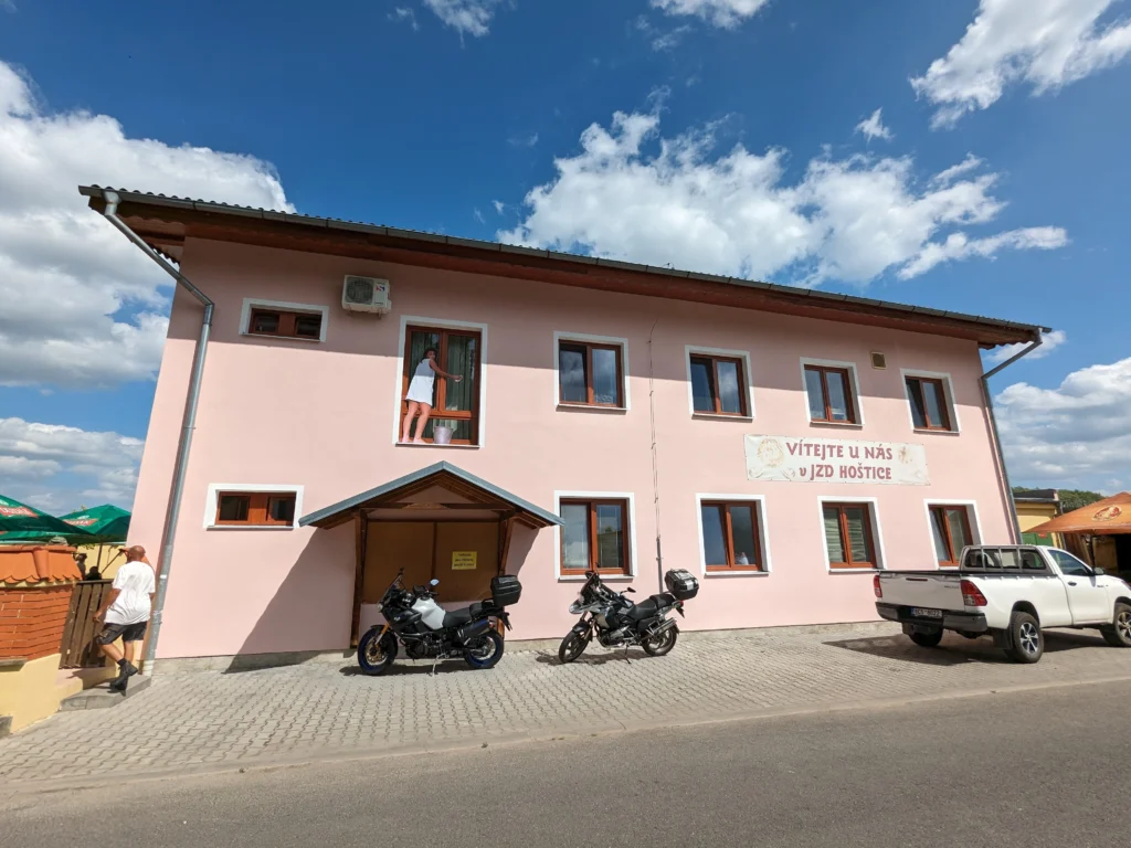 Růžová budova s nápisem Vítejte u nás v JZD Hoštice, v okně je nalepený plakát s ženou, která myje okna