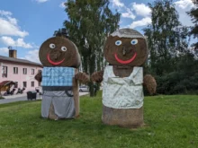 Dva velcí panáci ze slámy, jeden ztvárňuje pána, druhý paní, stojí na louce i růžového domu