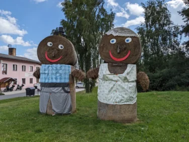 Dva velcí panáci ze slámy, jeden ztvárňuje pána, druhý paní, stojí na louce i růžového domu