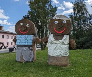 Dva velcí panáci ze slámy, jeden ztvárňuje pána, druhý paní, stojí na louce i růžového domu
