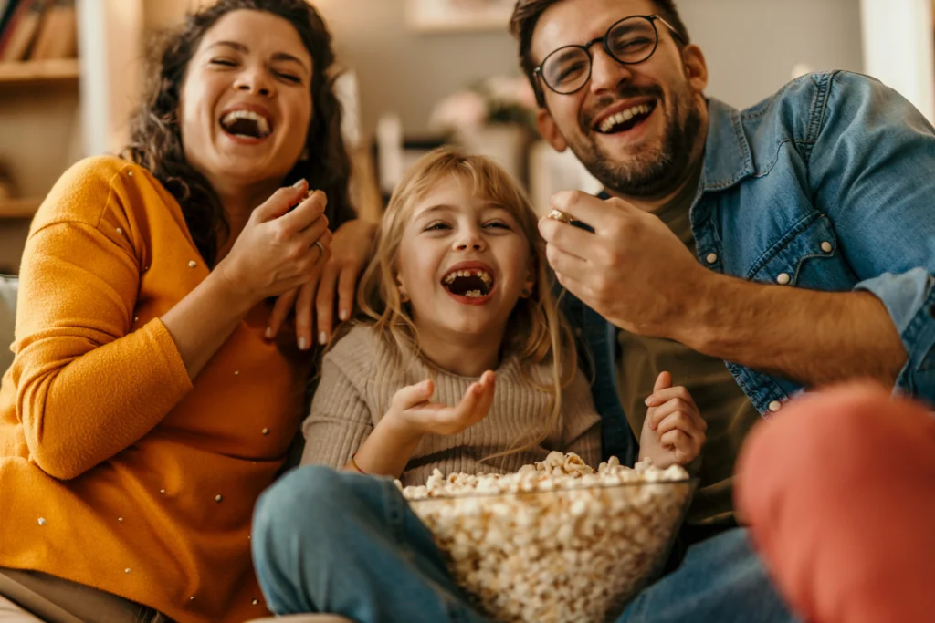 Rodina, táta, máma, holčička, sedí na gauči, jedí popcorn a smějí se, dívají se na televizi