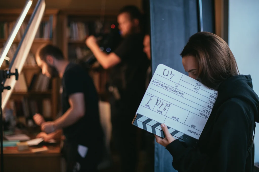 Fotografie z natáčení, je vidět kameraman, klapka a režisér, všude je zmatek