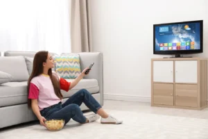 Žena sedí na zemi opřená o gauč a dívá se na televizi