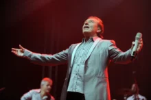 Zpěvák roztahuje paže k publiku a zpívá směrem vzhůru