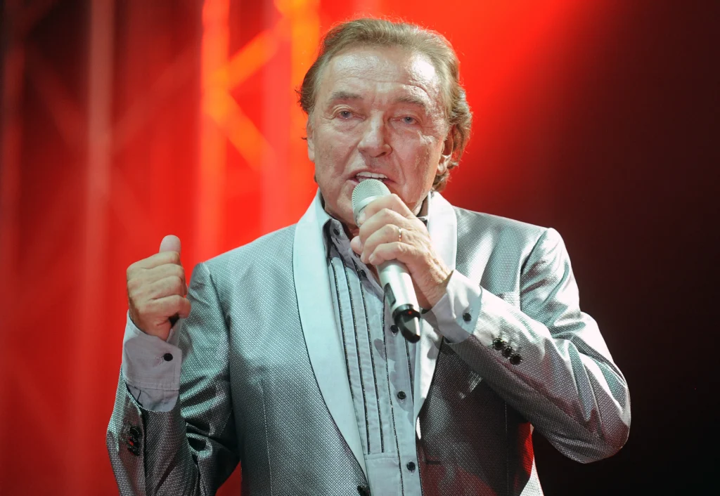 shutterstock_356238056 - VIPshow Zpěvák ve světlém obleku zpívá, za ním je červené pozadí. Je to Karel Gott