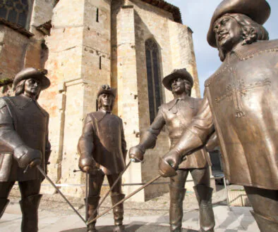 Sousoší čtyř mužů v kloboucích, pláštích a s kordy. Bronzové sochy u historické stavby z kamene.