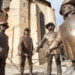Sousoší čtyř mužů v kloboucích, pláštích a s kordy. Bronzové sochy u historické stavby z kamene.