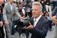 Muž se šedými vlasy v modrém obleku drží fotoaparát s velkým objektivem