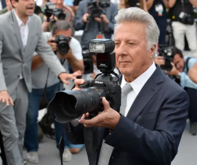 Muž se šedými vlasy v modrém obleku drží fotoaparát s velkým objektivem