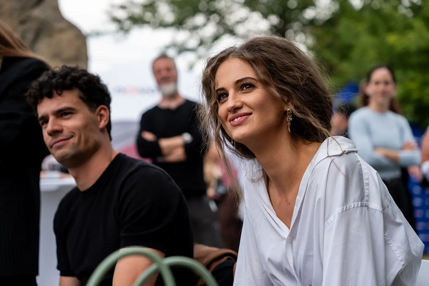 TV Nova_Boží plán_premiéra Špejchar 4 - VIPshow Žena s dlouhými vlasy a v bílé košili sedí venku a vedle ní, o kousek dál, sedí muž v černém tričku