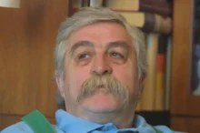 Starší muž s šedivými vlasy a šedivými vousy sedí v křesle a dívá se do strany