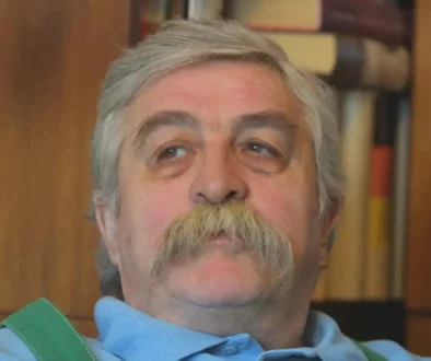 Starší muž s šedivými vlasy a šedivými vousy sedí v křesle a dívá se do strany