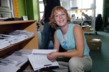 Žena v modrém tílku drží v ruce papíry a sedí `na bobku`