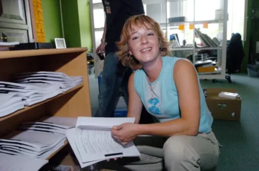 Žena v modrém tílku drží v ruce papíry a sedí `na bobku`