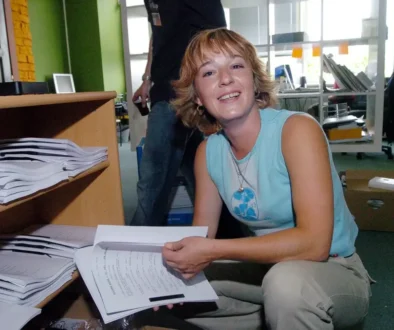 Žena v modrém tílku drží v ruce papíry a sedí `na bobku`