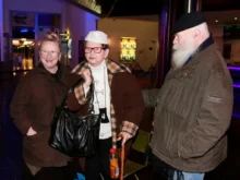 Three elderly friends in brown jackets chat together indoors, near a blue-lit wall.