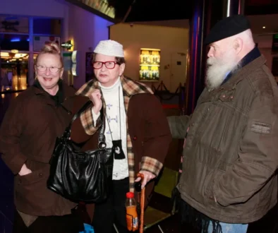 Three elderly friends in brown jackets chat together indoors, near a blue-lit wall.