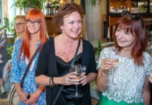 Three smiling women at a social event, holding wine glasses in a bar/restaurant setting.