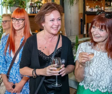 Three smiling women at a social event, holding wine glasses in a bar/restaurant setting.