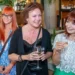 Three smiling women at a social event, holding wine glasses in a bar/restaurant setting.