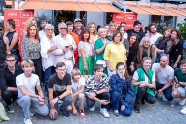 Velká skupina lidí, herců, za nimi jsou obchůdky a nápisy Ulice