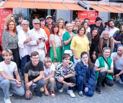 Velká skupina lidí, herců, za nimi jsou obchůdky a nápisy Ulice