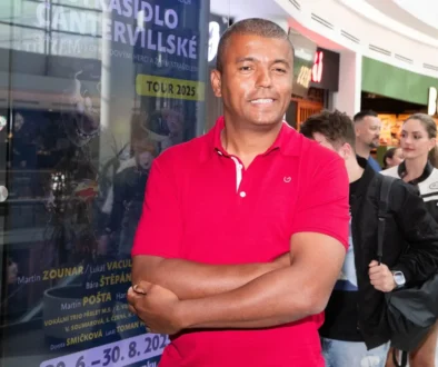 Muž tmavé pleti ve výrazně růžovém tričku stojí v nákupním centru a má ruce zkřížené na prsou