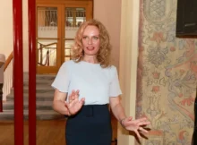 Žena v bílém tričku a tmavé sukni gestikuluje ve vestibulu