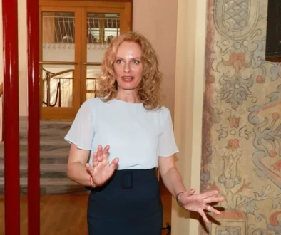 Žena v bílém tričku a tmavé sukni gestikuluje ve vestibulu