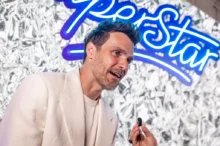 Muž v bílém oblečení se usmívá a mluví do mikrofonu, za ním je poutač Superstar