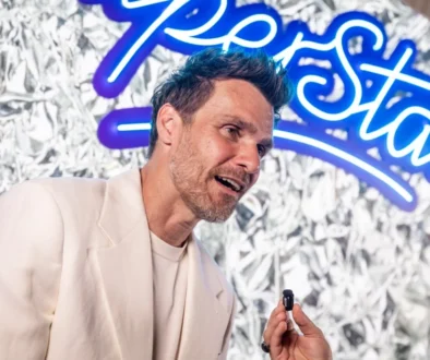 Muž v bílém oblečení se usmívá a mluví do mikrofonu, za ním je poutač Superstar
